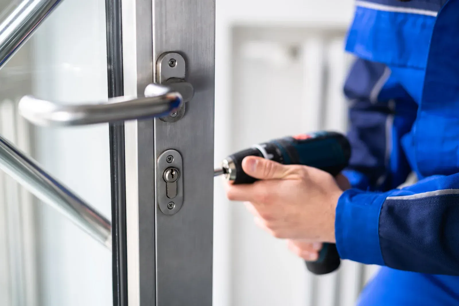 Person in blue overalls using a drill on a metal door with a handle and lock.