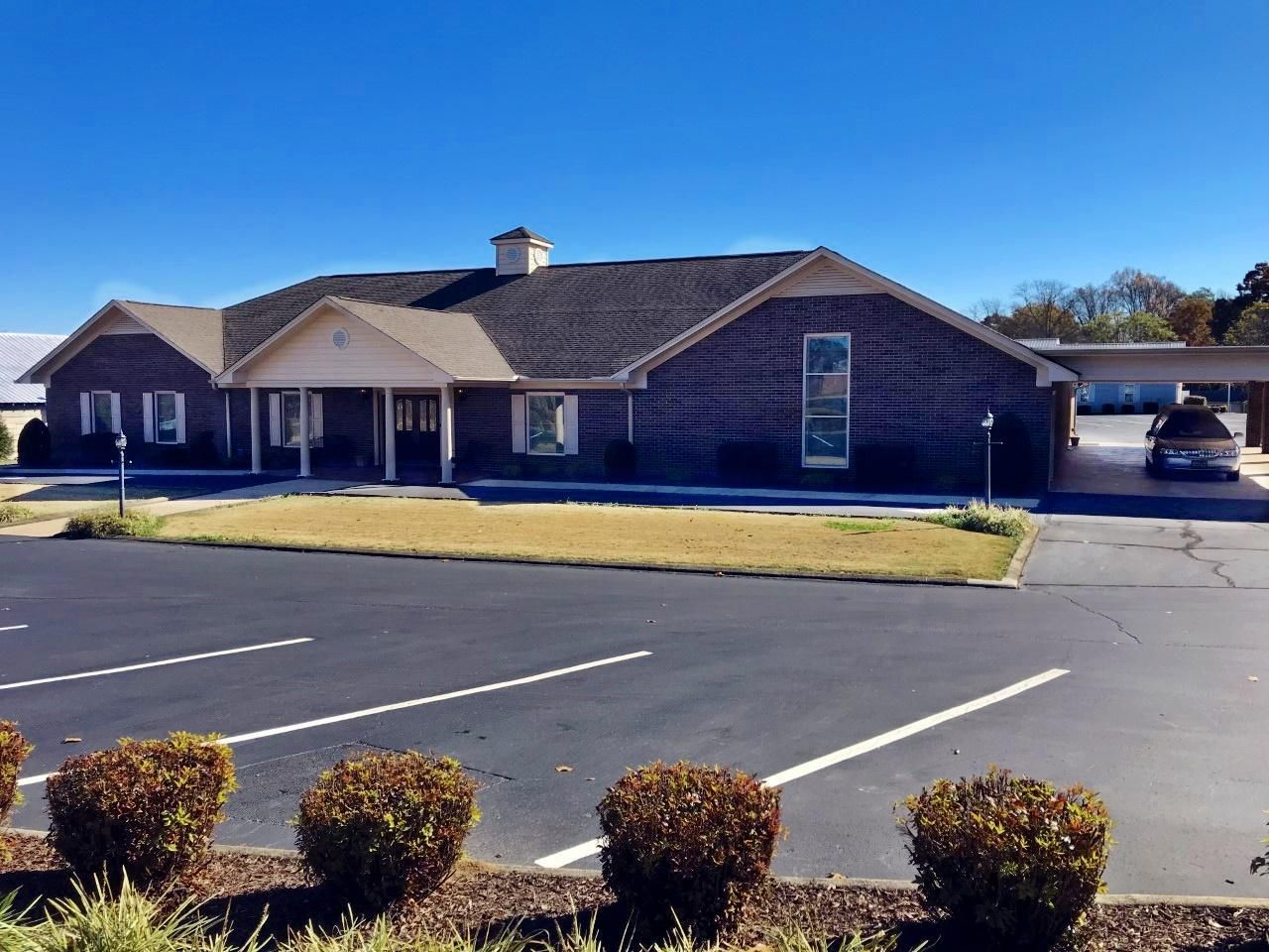 Brick building with dark roof, parking lot, and bushes in front on a sunny day.