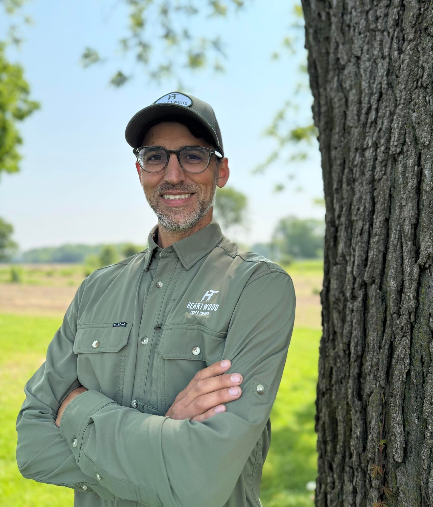 Man in green shirt and hat leaning against a tree, arms crossed, smiling outdoors.