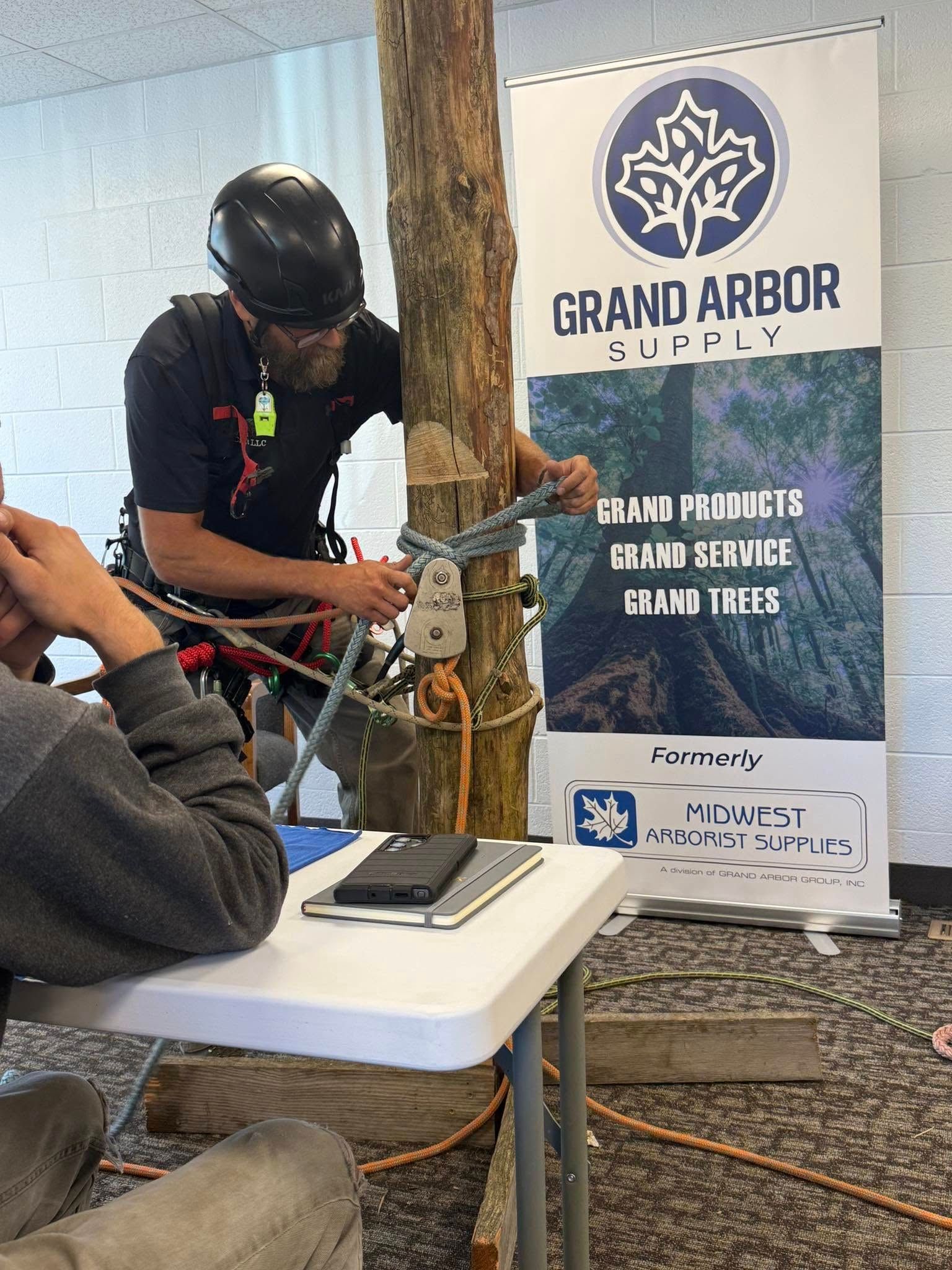 Arborist tying a knot on a tree trunk during a demonstration, in front of a Grand Arbor Supply banner.