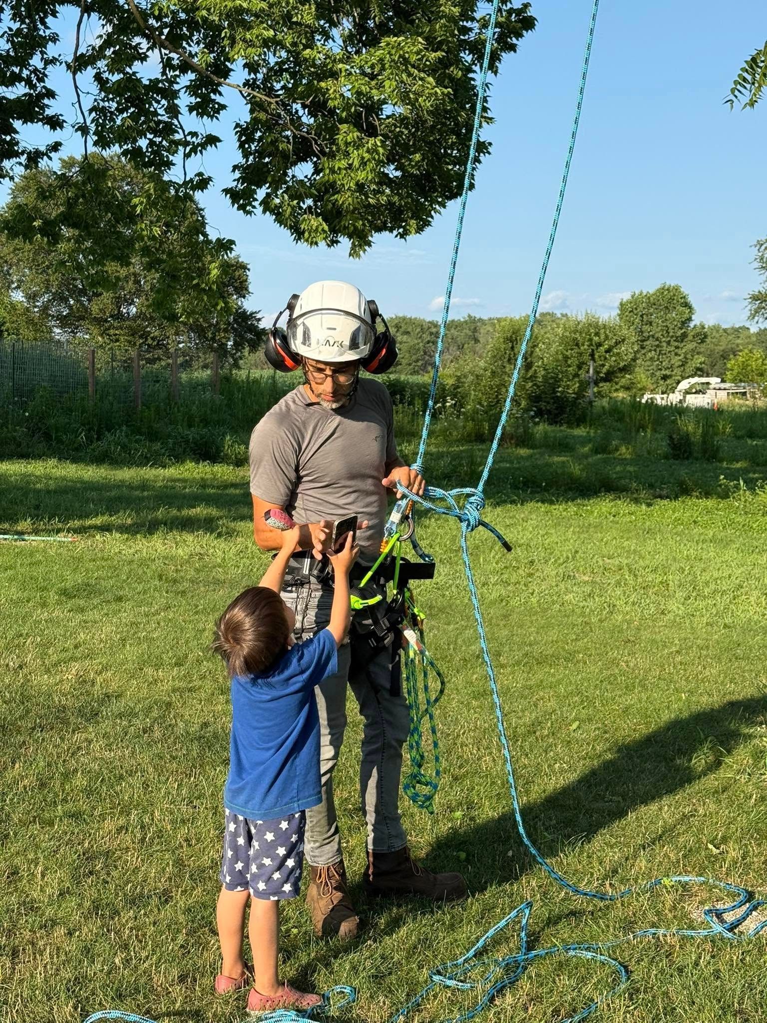 Person in safety gear with a young child outdoors, touching a climbing rope. Green grass, trees in background.