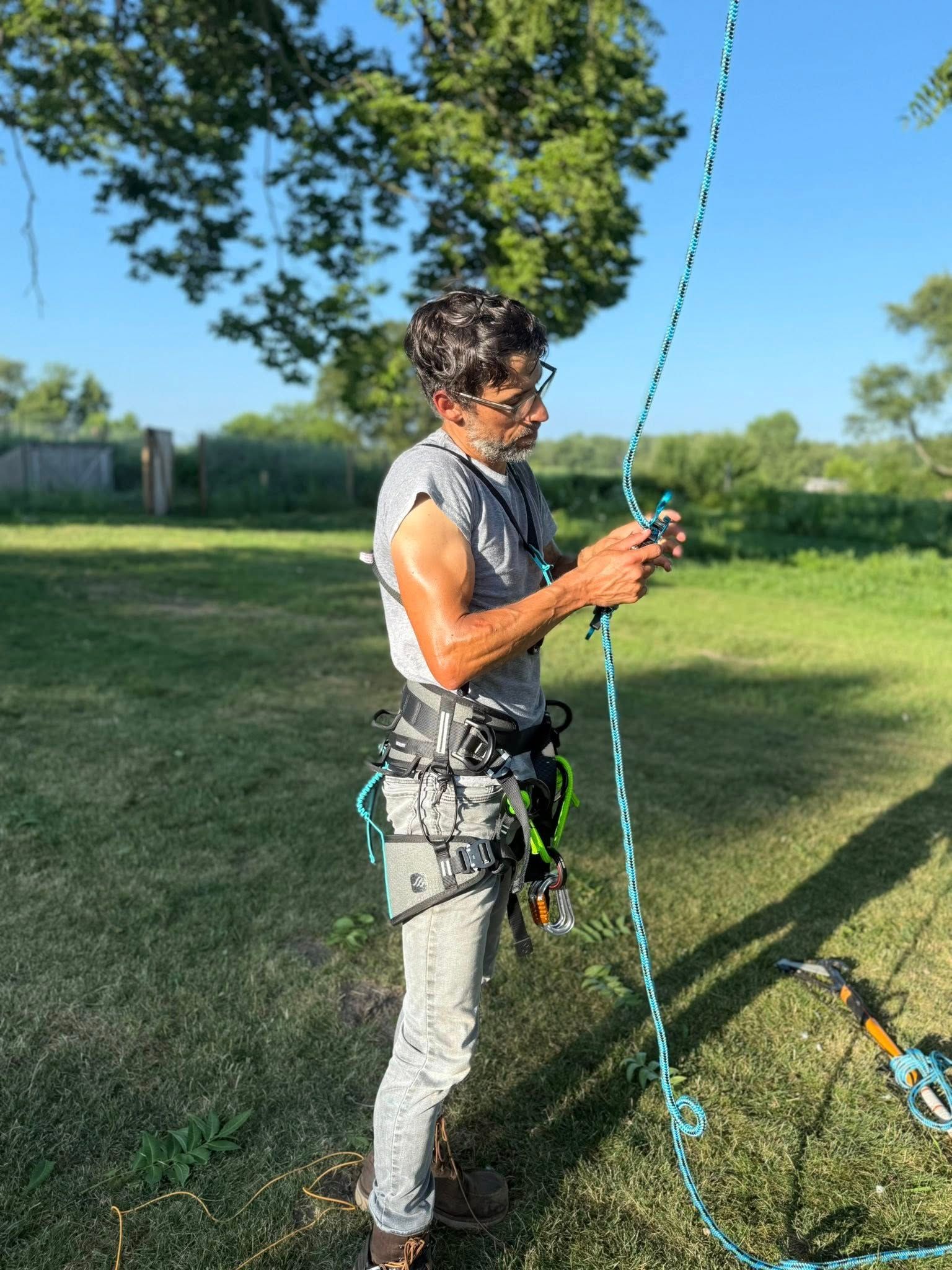 Person in climbing gear, holding a rope outdoors, prepping to climb a tree. Green grass, blue rope.