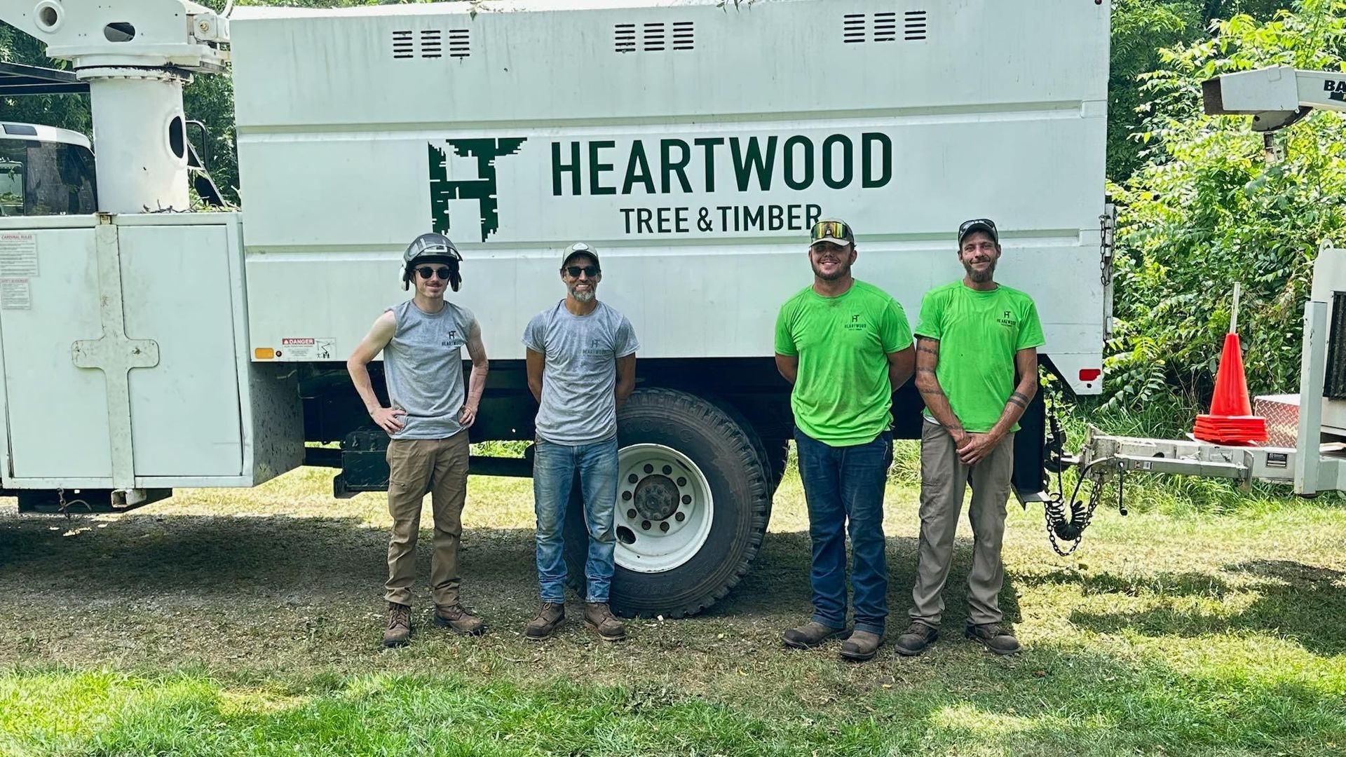 Four workers pose in front of a Heartwood Tree & Timber truck. They stand on grass, wearing work clothes.