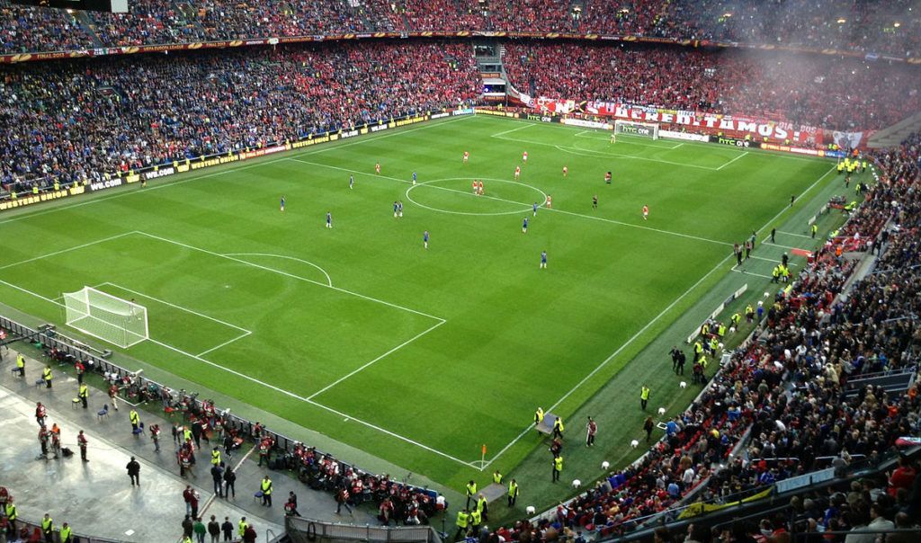 Een voetbalwedstrijd in een volgepakt stadion, spelers op een felgroen veld met fans op de tribunes.