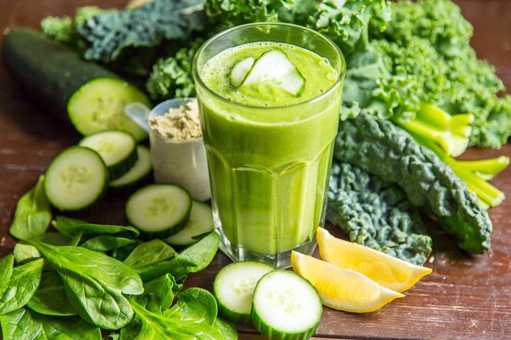 Groene smoothie in een glas, omringd door boerenkool, spinazie, komkommer en citroenpartjes.