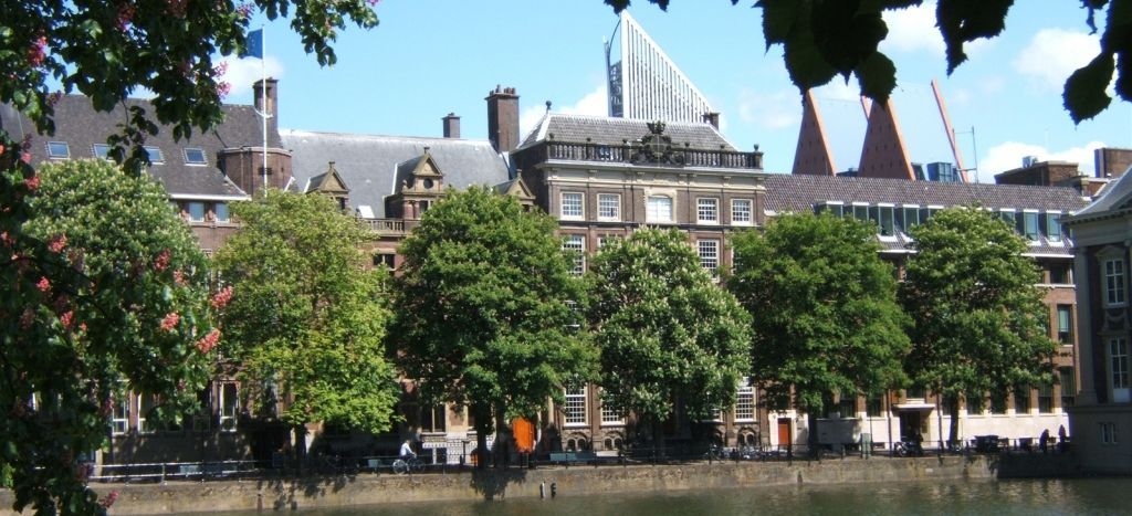 Een met bomen omzoomde rivieroever met historische gebouwen en daken aan de overkant, onder een heldere hemel.
