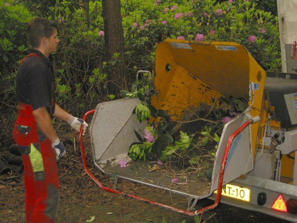 Een arbeider staat naast een beschadigde oranje machine in een tuin, met gebroken beton en verspreid puin.
