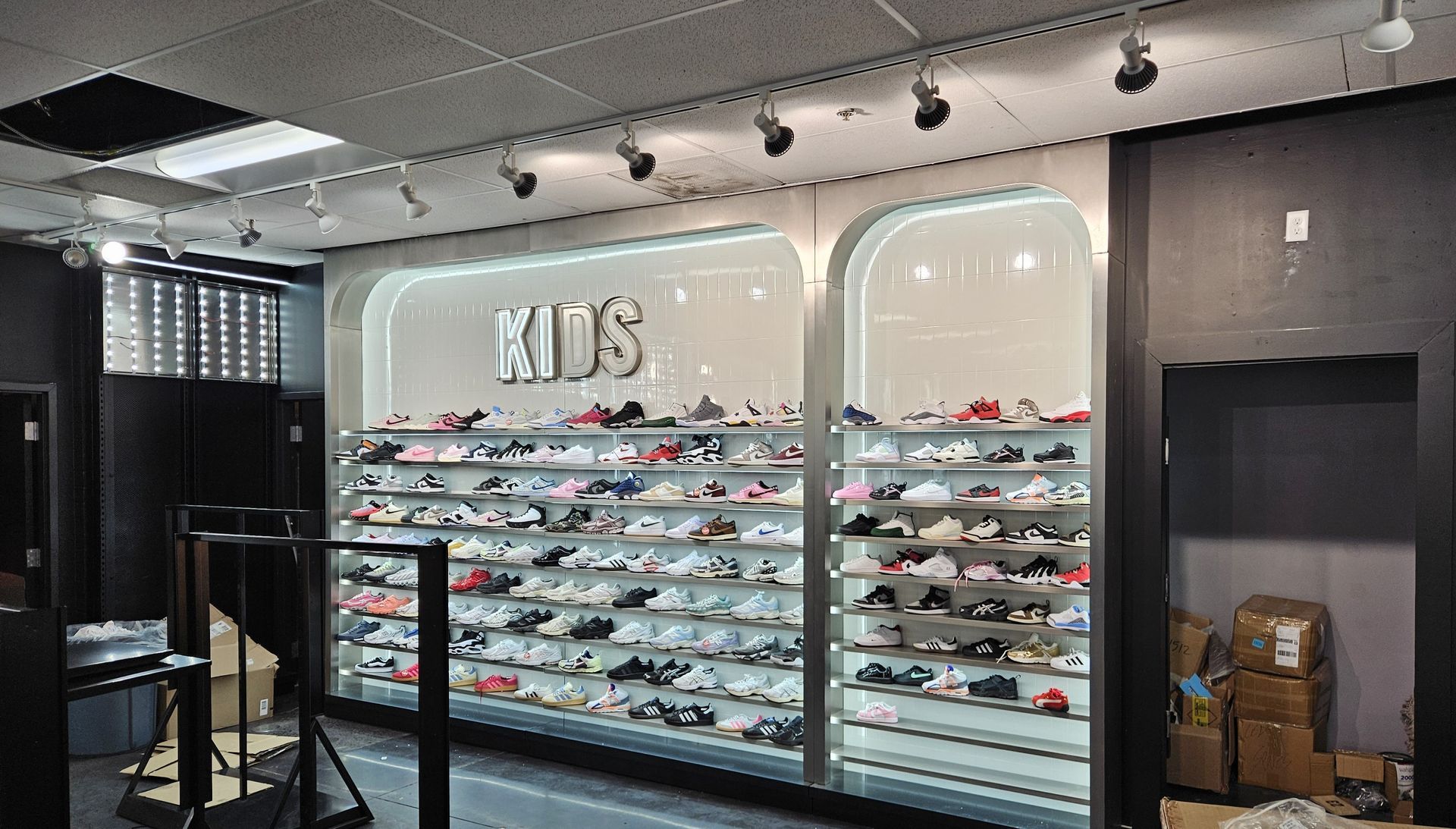 Shoe store display with rows of sneakers under a large “KIDS” sign and track lighting