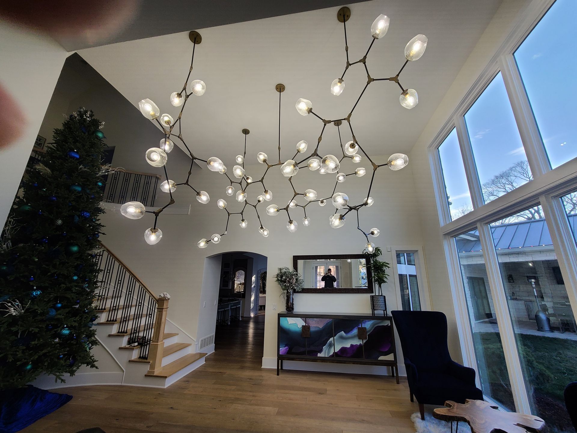 Bright modern living room with sculptural chandeliers, staircase, and large windows overlooking a patio