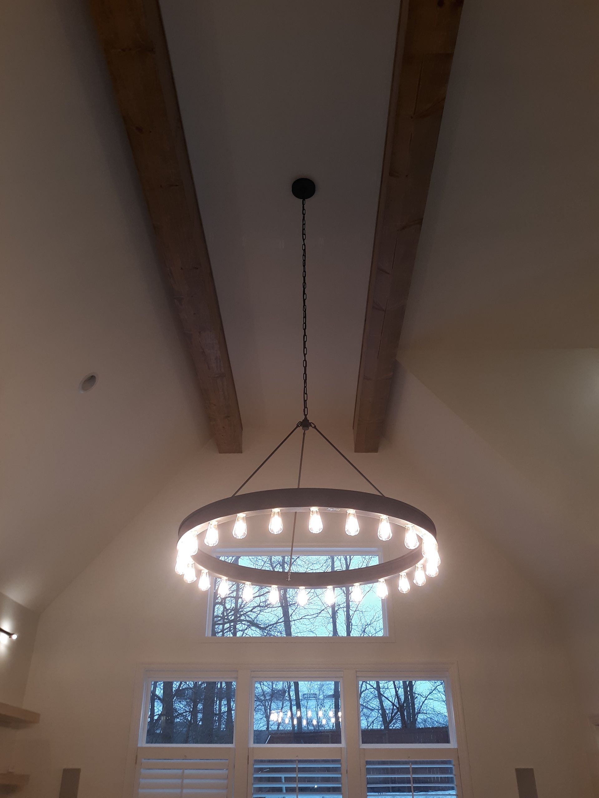 Large circular chandelier hanging from a high vaulted ceiling above tall windows and wooden beams