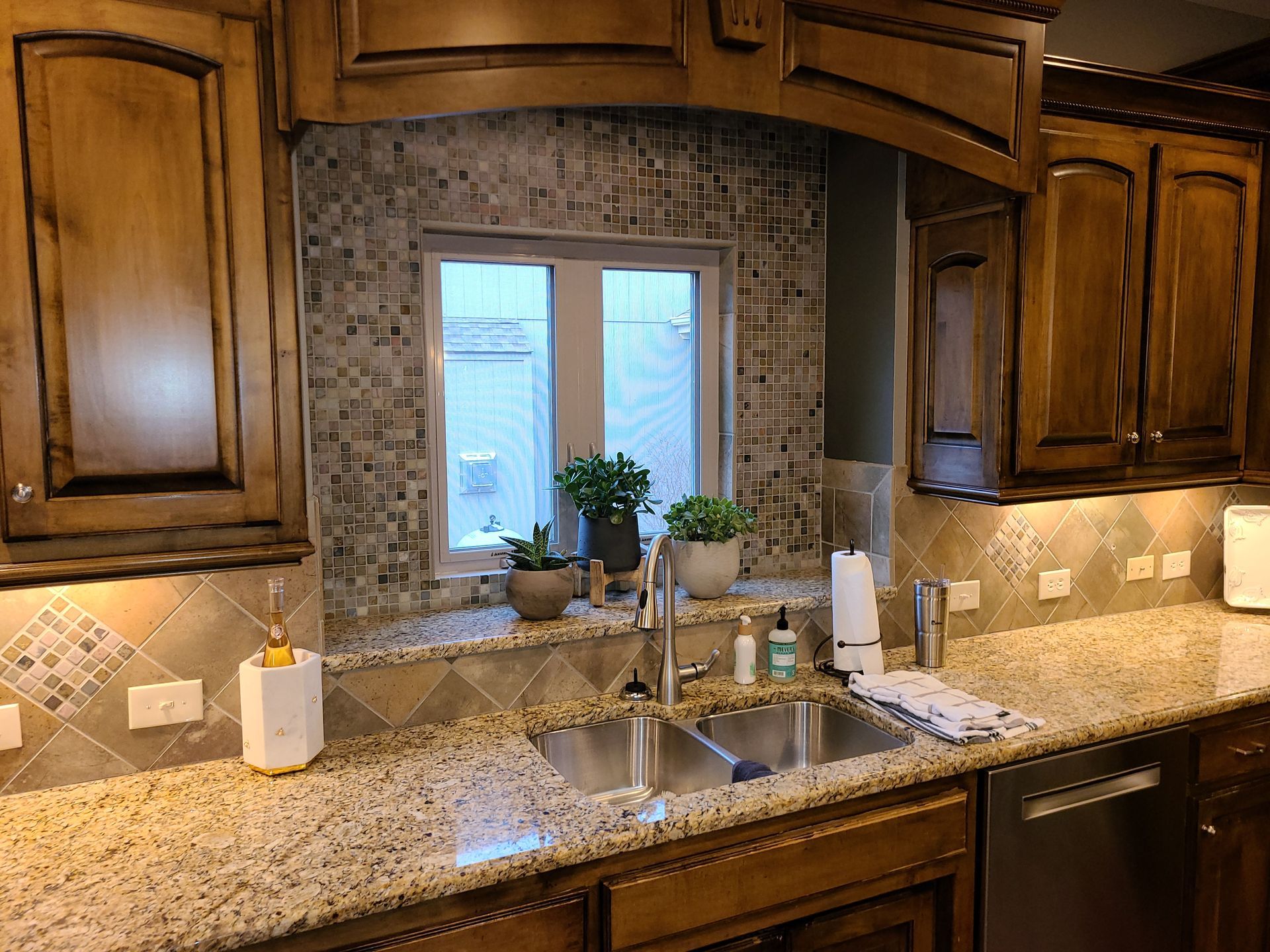 Kitchen sink beneath window, framed by wood cabinets and granite countertops with mosaic tile backsplash.