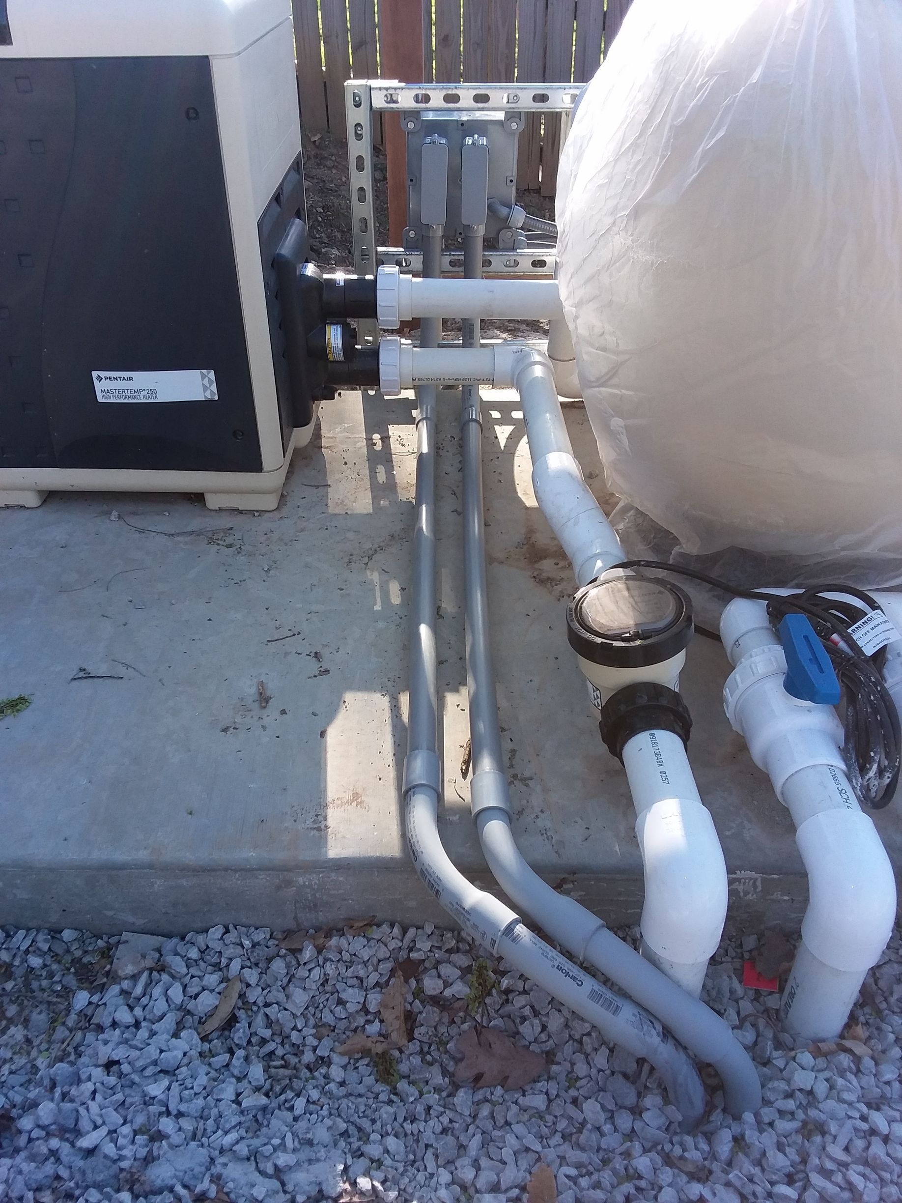 Outdoor HVAC equipment with pipes, valves, and concrete pad beside gravel and a large tank