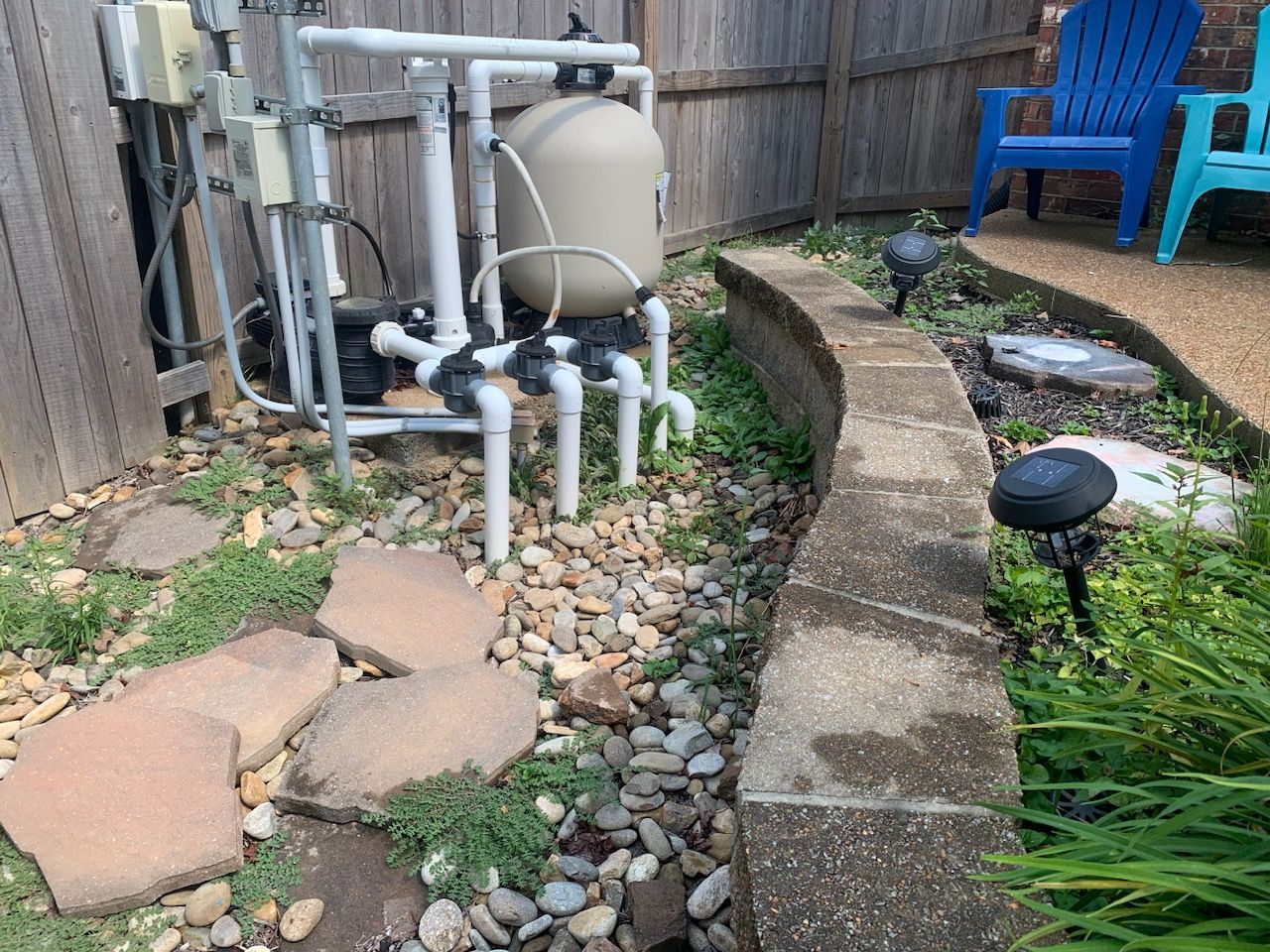 Pool equipment by a stone wall and garden bed with pebbles and greenery.