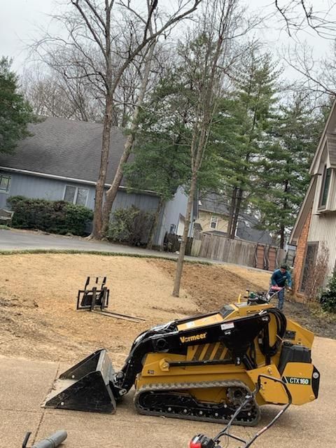 Yellow mini-skid steer on asphalt, operator using controls; residential setting with houses and trees.
