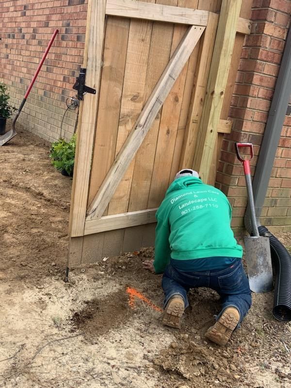 Man in green hoodie kneeling, working on wooden gate, next to a brick building and dirt area.