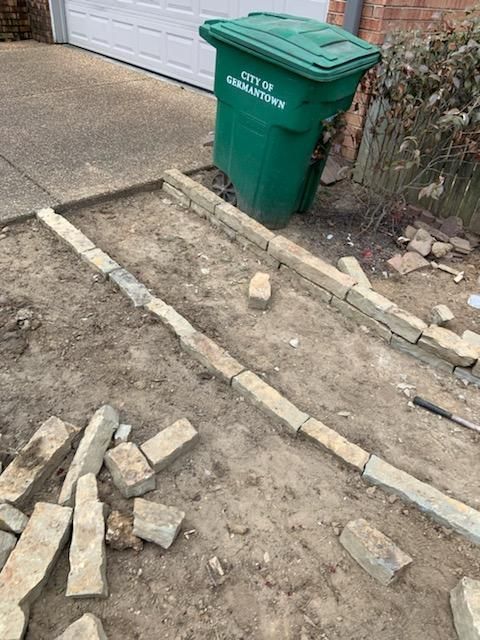 Construction of a stone border along a driveway; green trash can nearby; dirt and stone blocks visible.