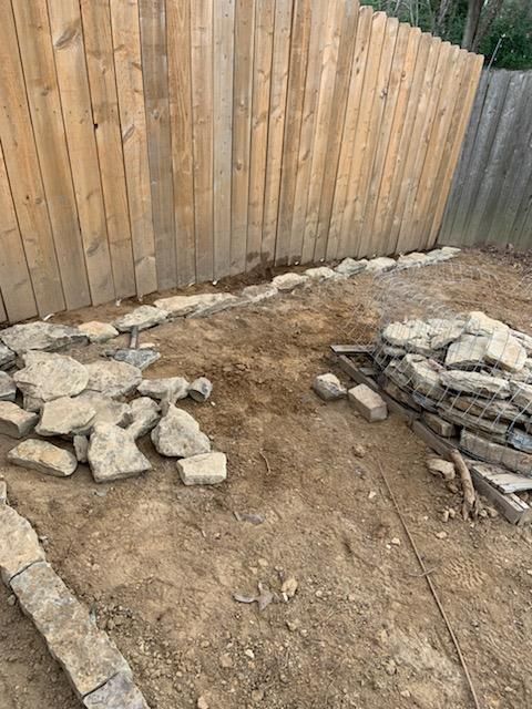 A backyard with a wood fence, dirt ground, and stone edging along the fence. Stones are in piles and placed on the ground.