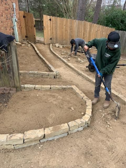 Two workers laying stone edging in a yard. One rakes dirt, the other works on a path.