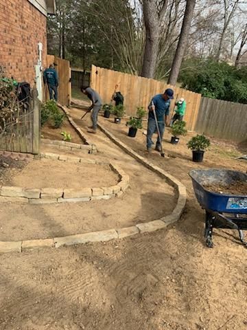 People building a garden path with stone borders. Shovels and plants visible.
