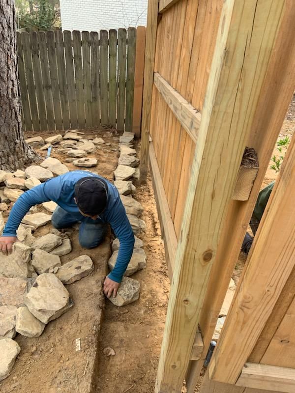 Man kneeling, arranging stones along a dirt border next to a wooden fence in a backyard.