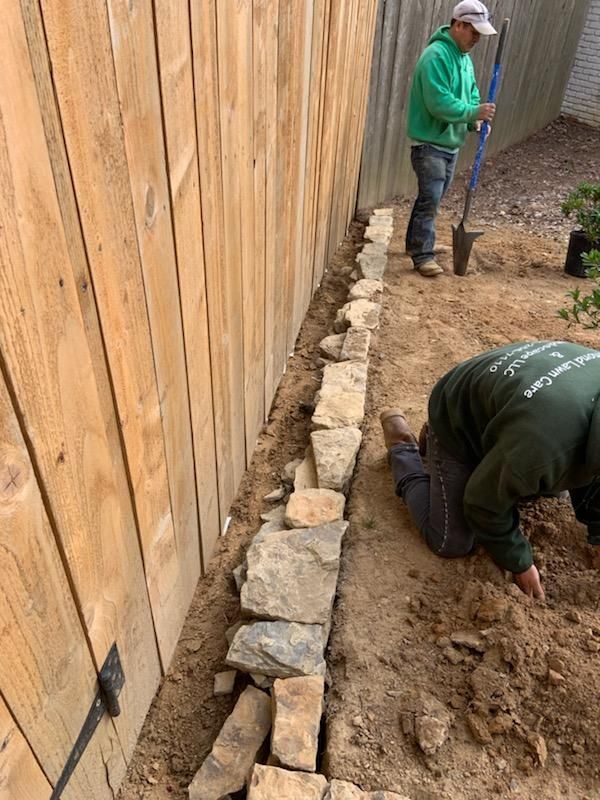 Two people building a stone border along a wooden fence. One digs, the other places stones.