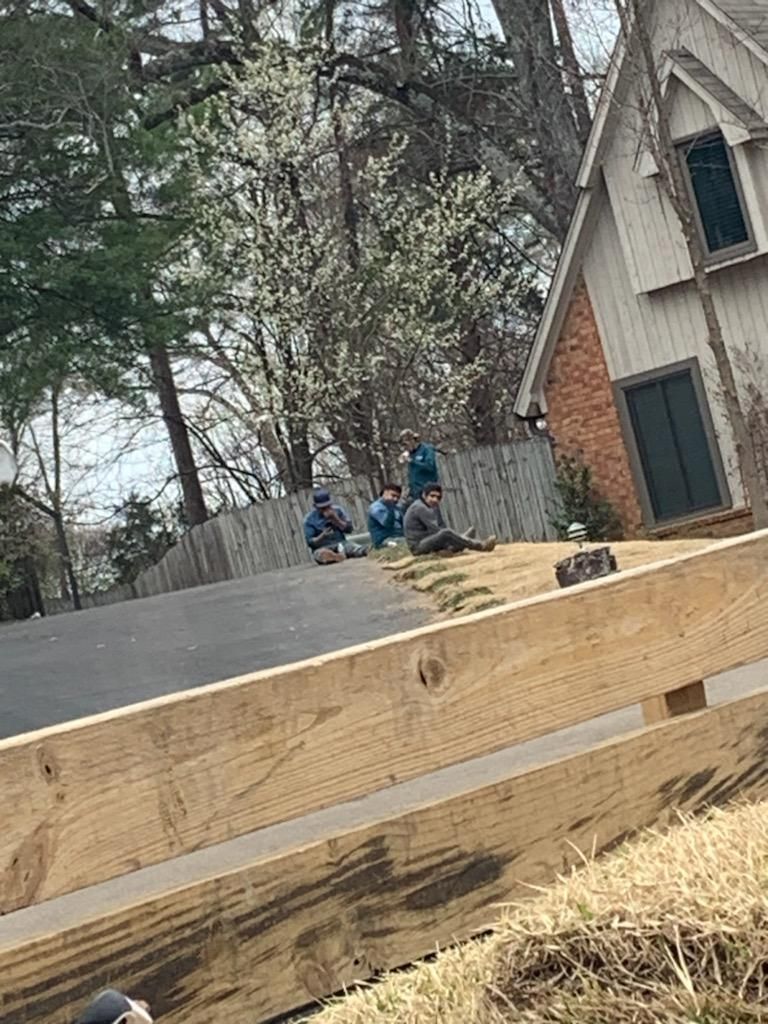 People on driveway near a house, sitting. Wooden fence in foreground. Trees, spring.
