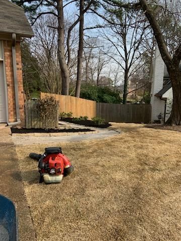 A red leaf blower sits on dry grass in a yard near a fence and house.