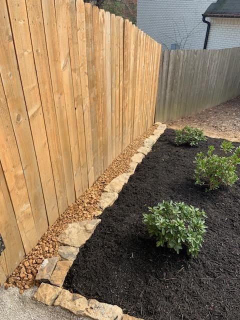 Wooden fence borders a garden bed with black mulch and small plants, edged with tan stones.