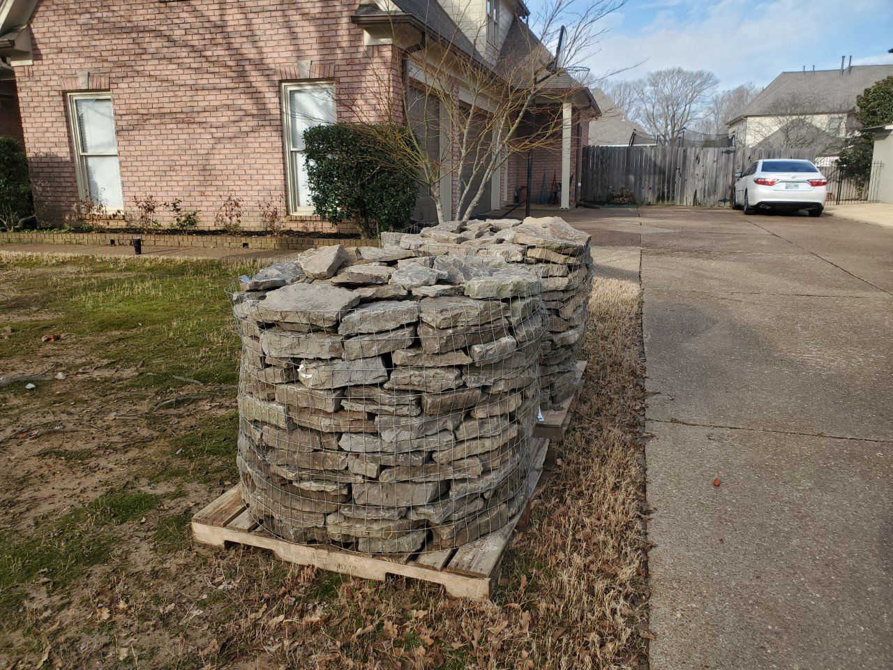 Stacked stone on a pallet sits near a driveway in front of a brick house. A car is parked in the background.