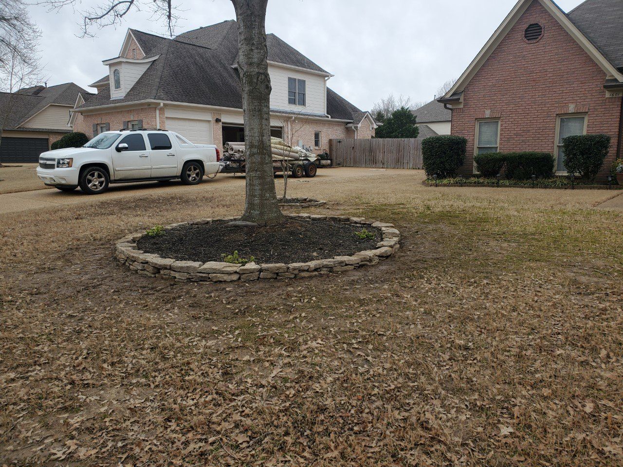 A white truck parked in front of a two-story house with brown grass, tree in focus with a rock border.
