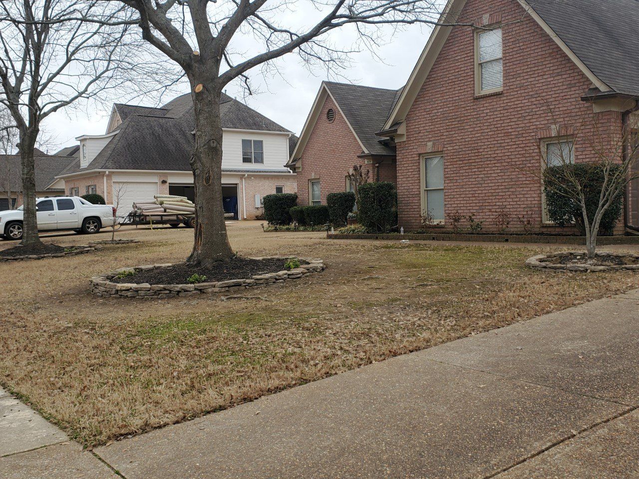 Residential street scene with houses and bare trees; lawn with brown grass and a sidewalk in the foreground.