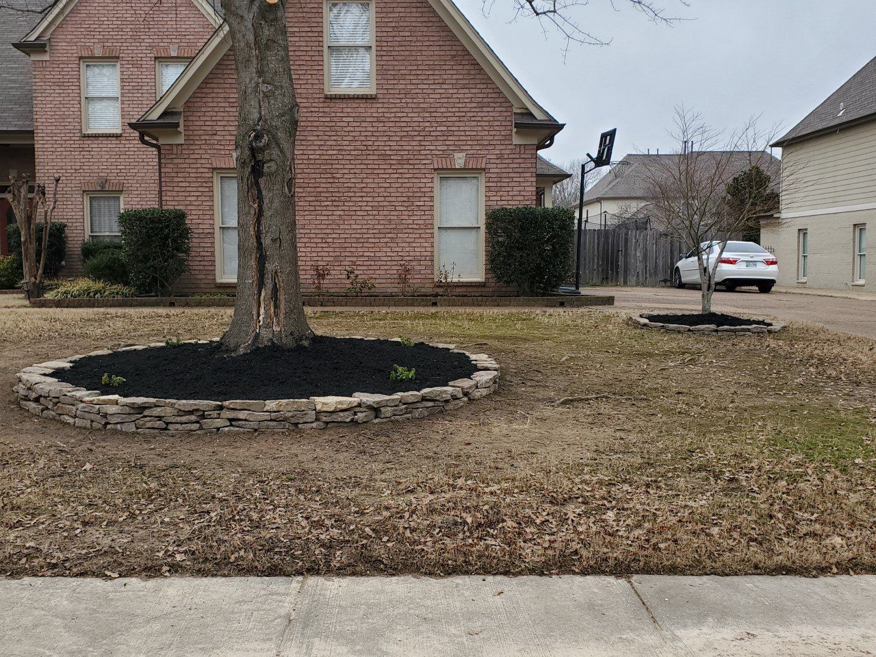 Brick house with tree in front yard, surrounded by stone and black mulch. Brown grass, overcast sky.
