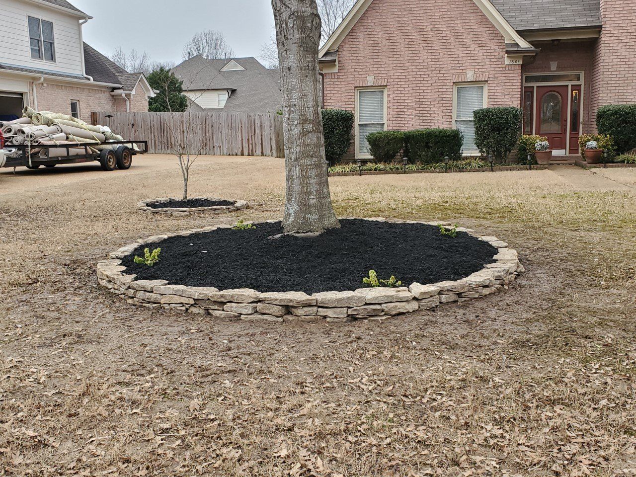 Circular tree bed with dark mulch, surrounded by a stone border, in front of a brick house on a lawn.