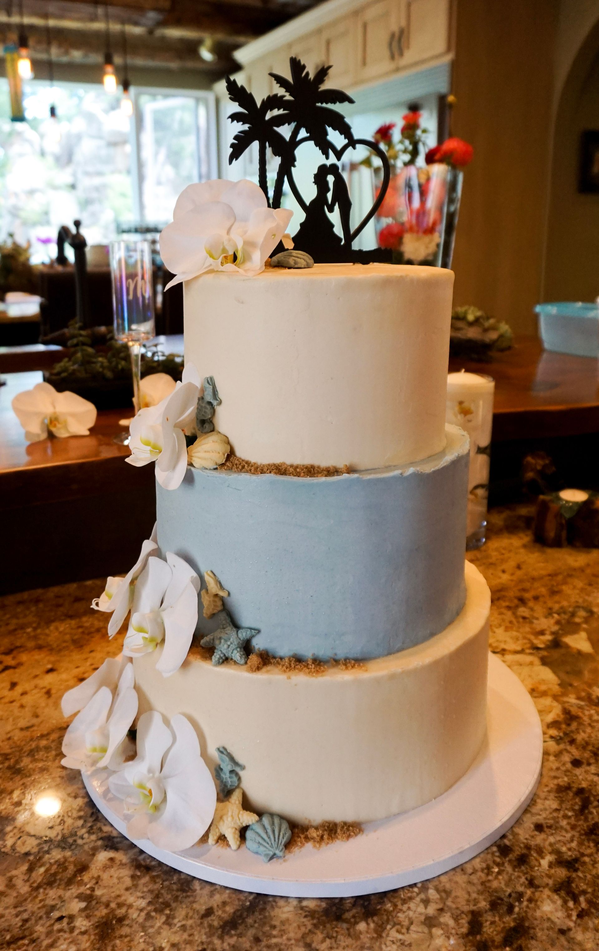 Three-tier white and blue wedding cake with flowers and palm tree topper on a table