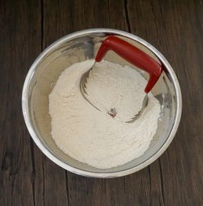 A metal bowl of white flour with a red-handled sifter on a dark wooden surface.