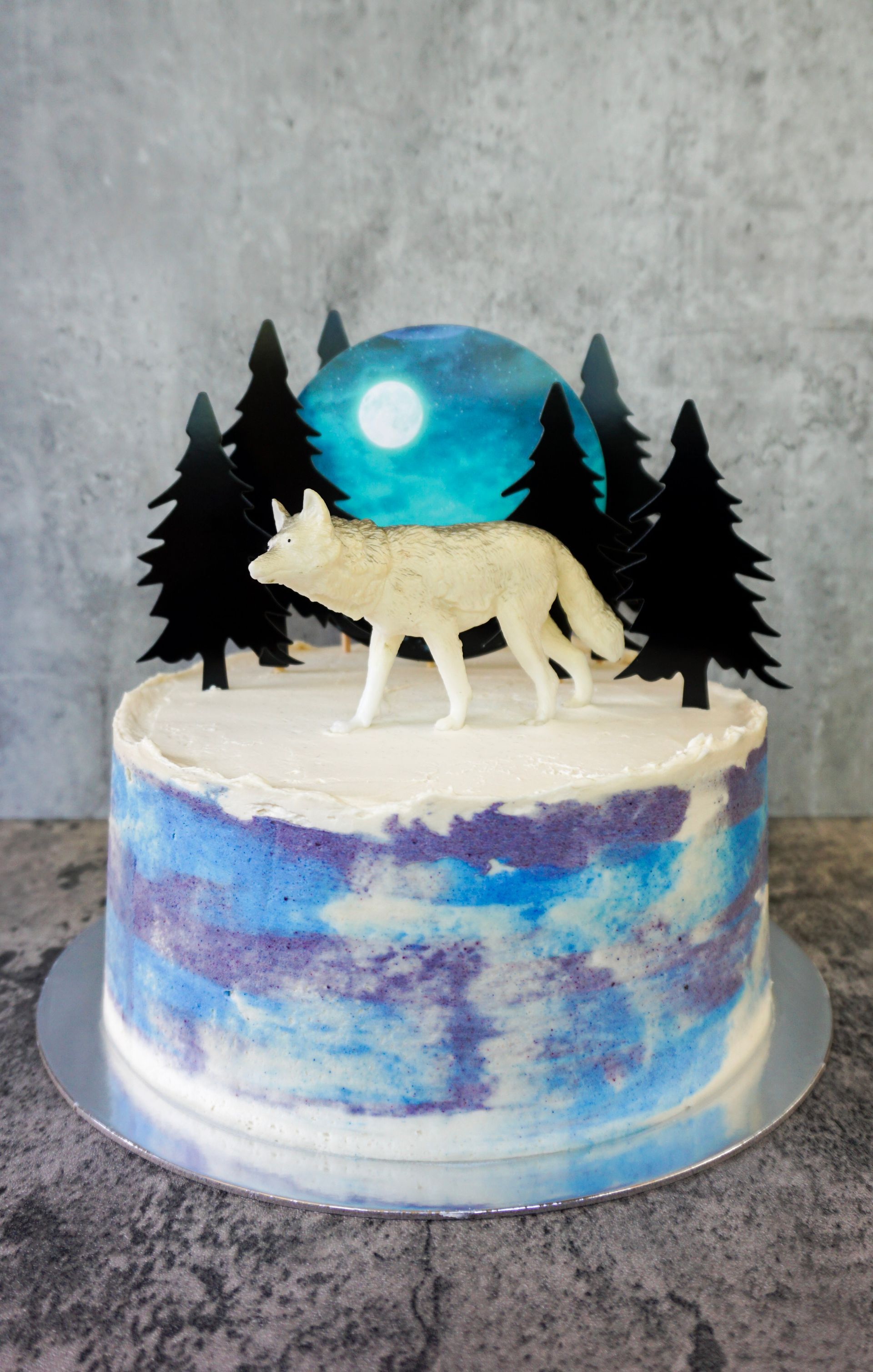 Winter-themed cake with blue watercolor frosting, white wolf figurine, pine trees, and a moonlit sky topper