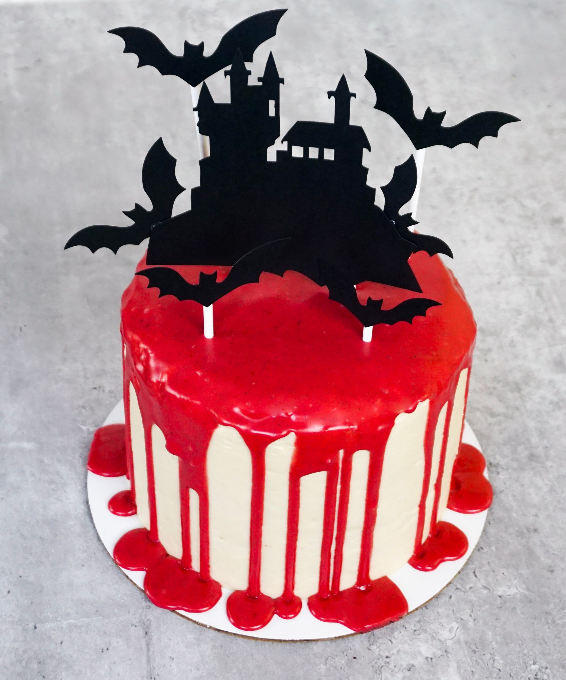 Red cake with white drips and black haunted house, bats, and cat Halloween topper