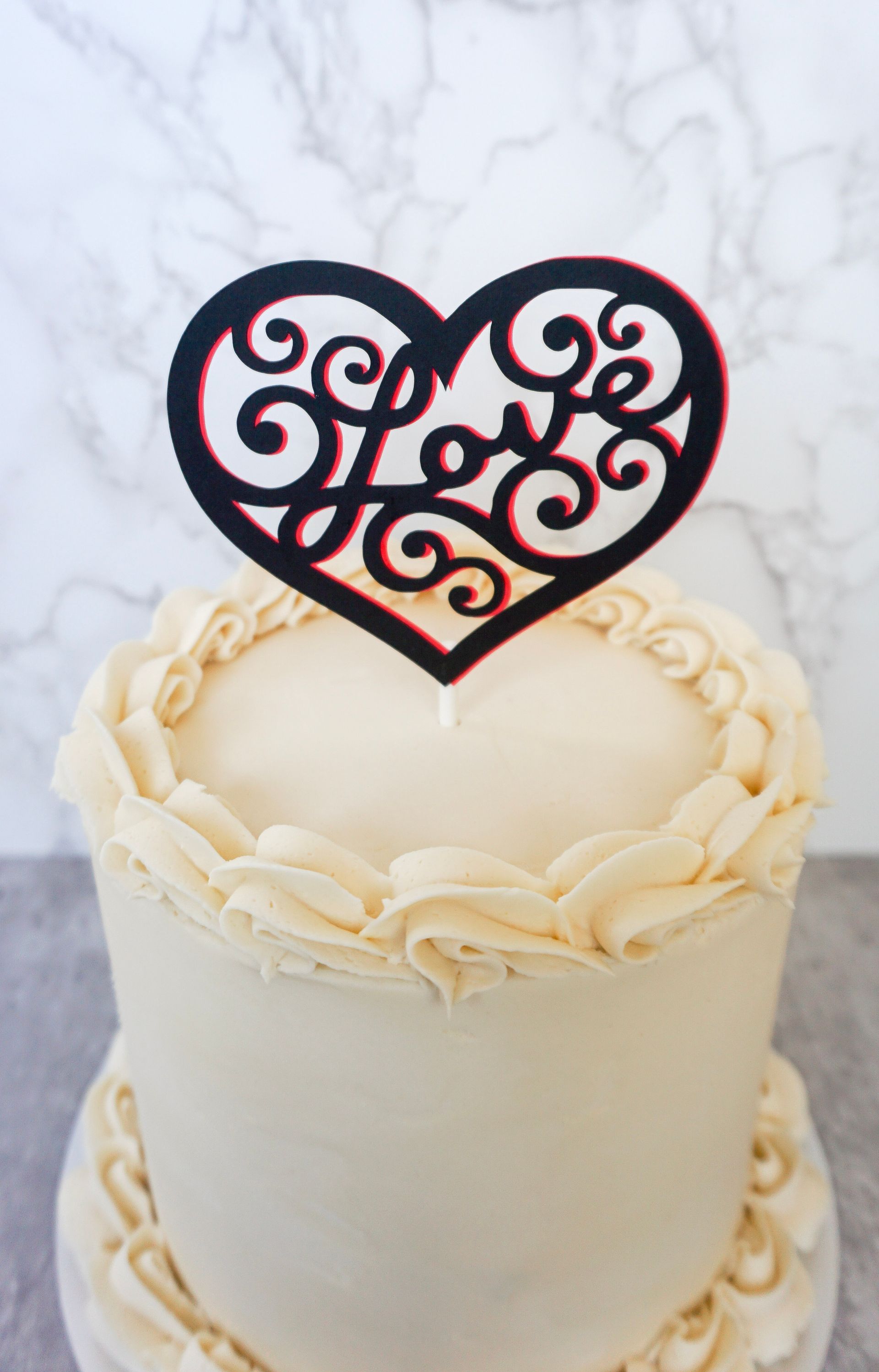 White frosted cake with a black filigree heart topper on a marble background