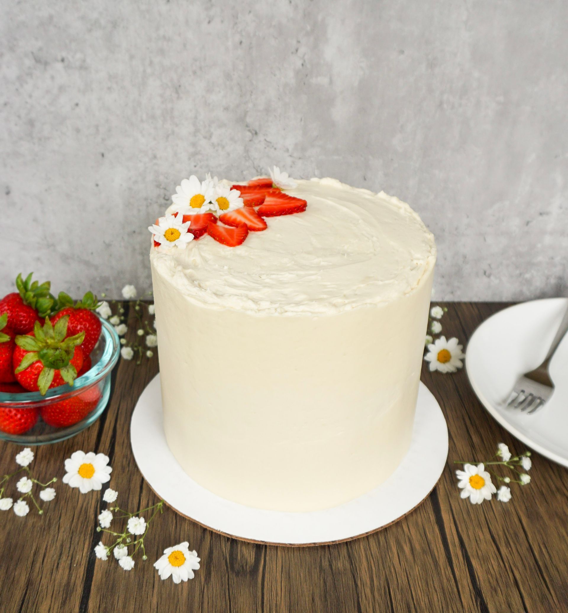 White frosted cake with strawberries and daisies on a rustic table, with strawberries and a plate nearby