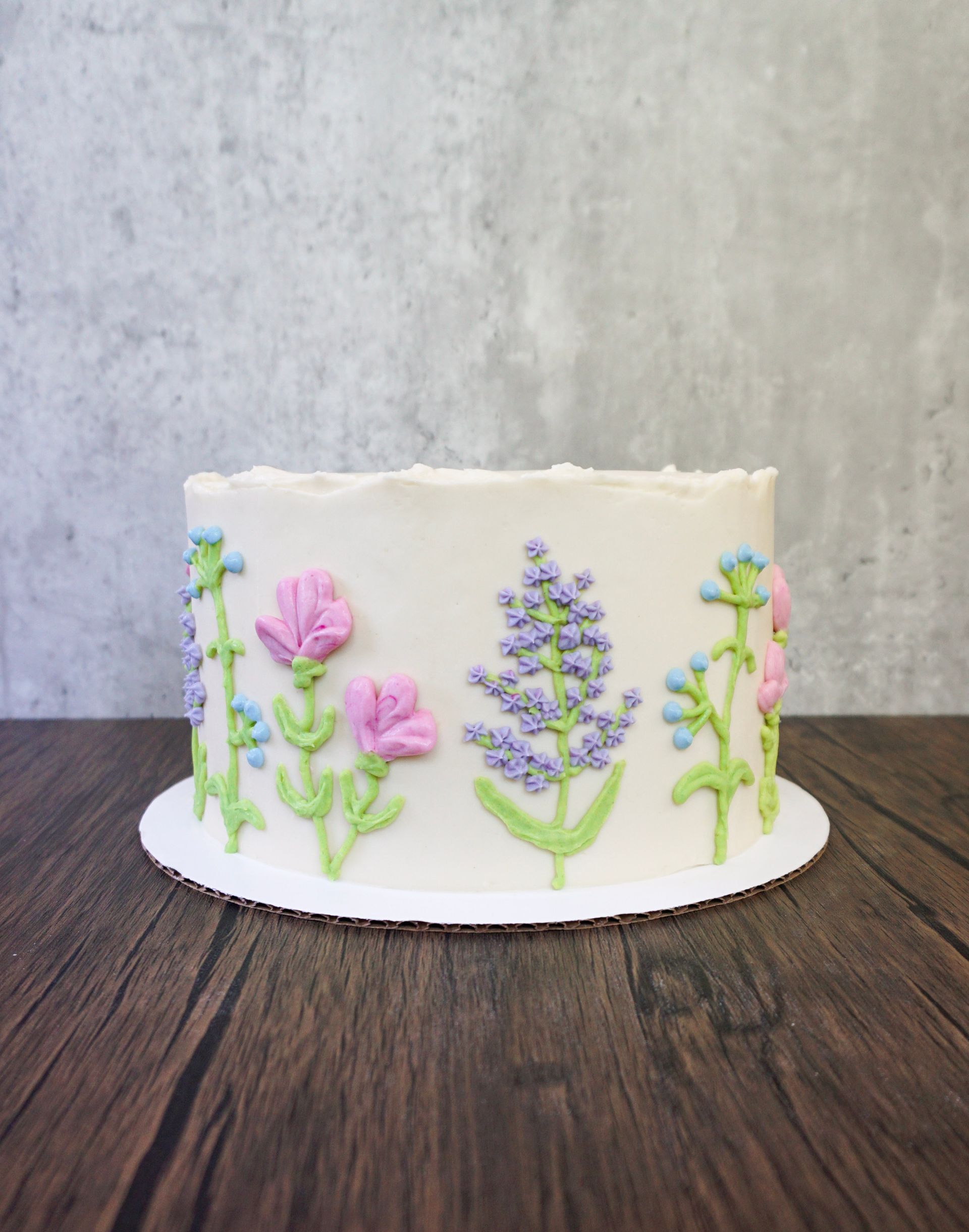 White cake decorated with pastel purple, pink, and green floral icing on a cake board