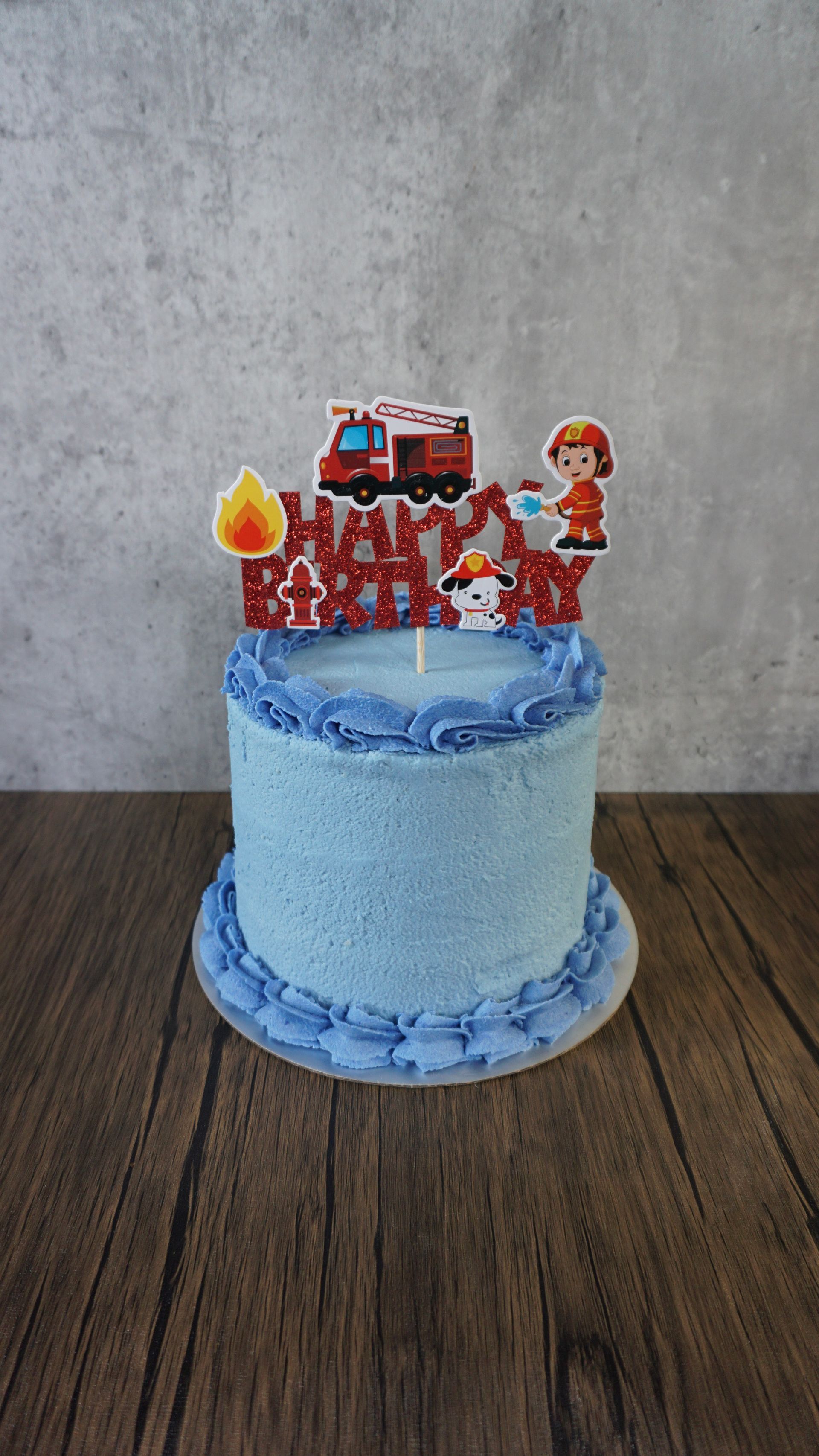 Light blue frosted cake with red fire truck and fire-themed topper on a wooden table