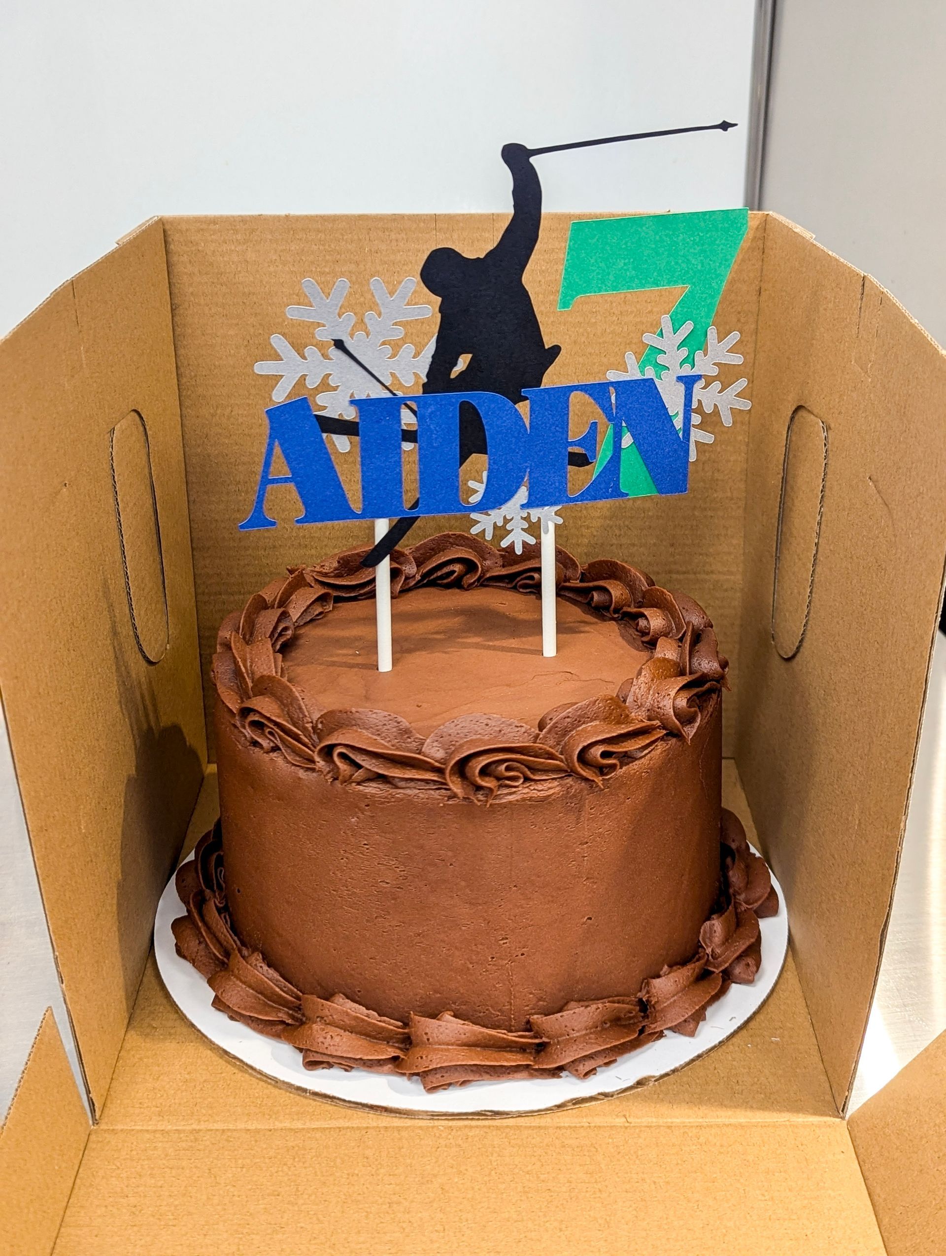 A chocolate birthday cake with an “AIDEN” topper and black cat silhouette inside a cardboard box.