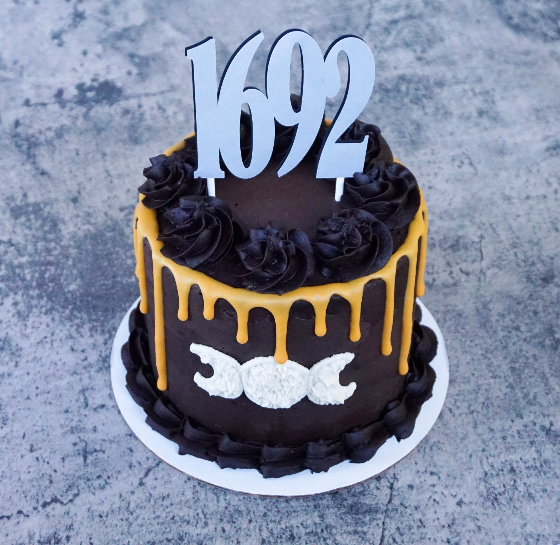 Black cake with gold drip, dark roses, and a silver “1692” topper on a white plate.