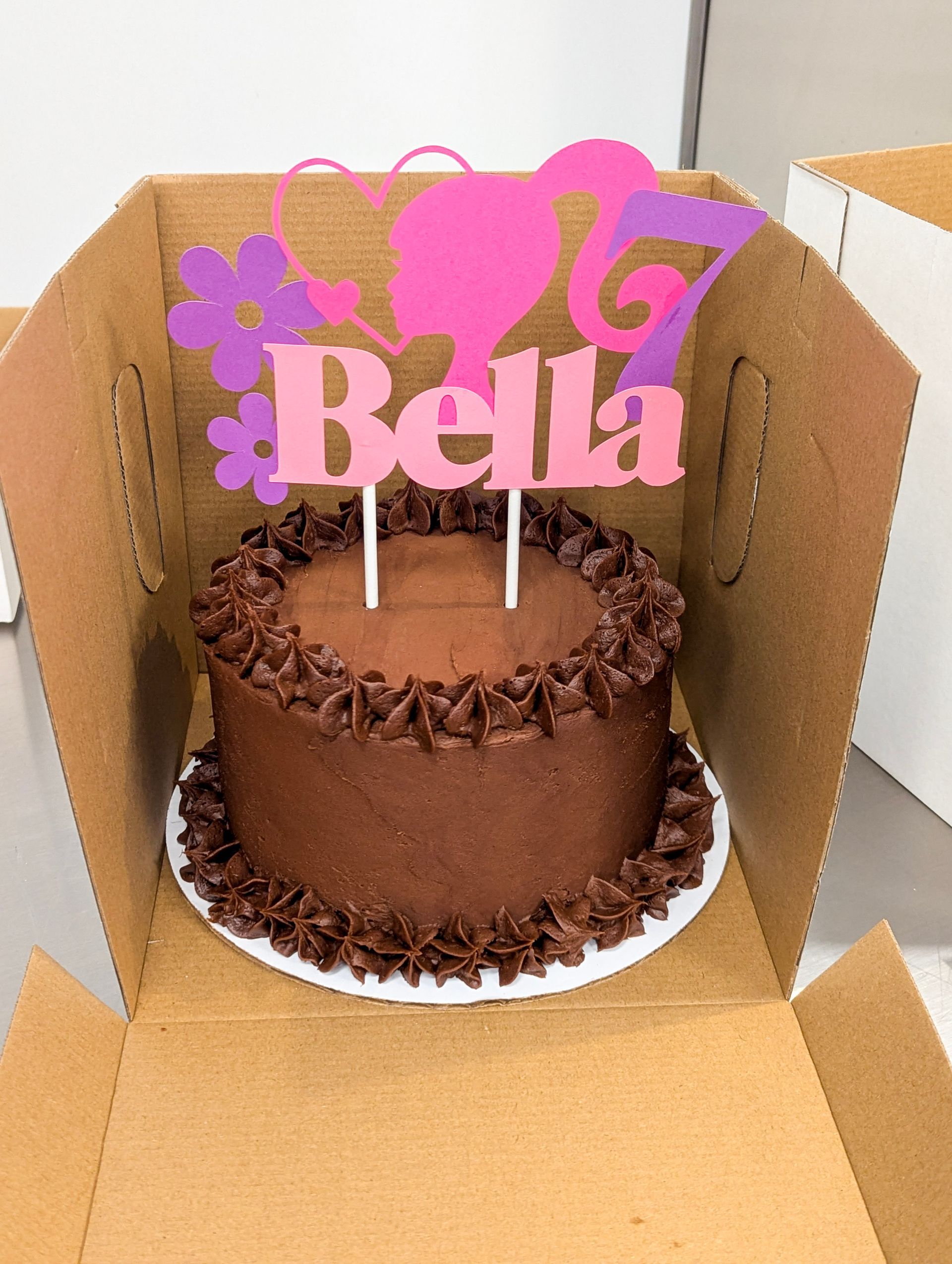 Chocolate cake with pink “Bella” topper inside an open cardboard cake box