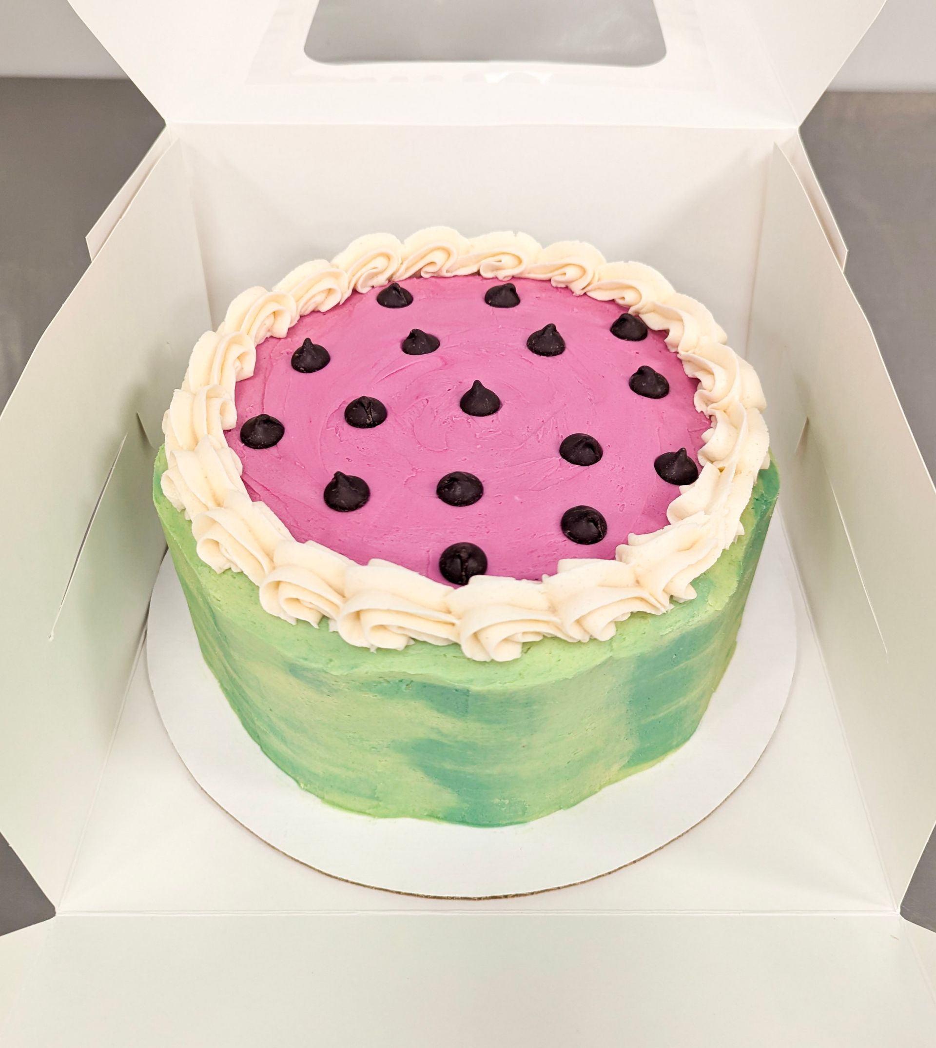 Round cake with pink top, black dots, and green-and-yellow sides in a white box