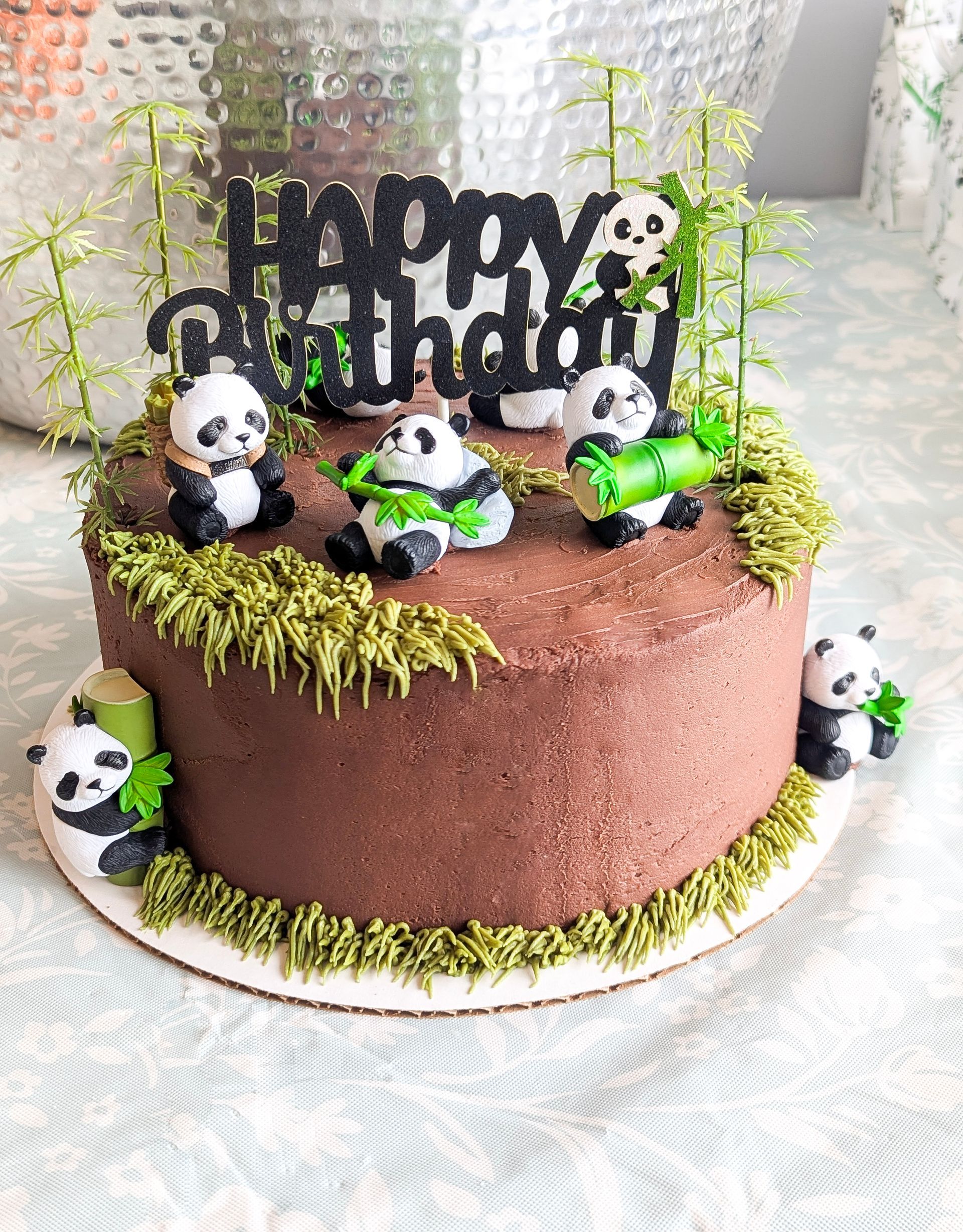 Brown birthday cake topped with panda figurines, green grass trim, and “Happy Birthday” topper