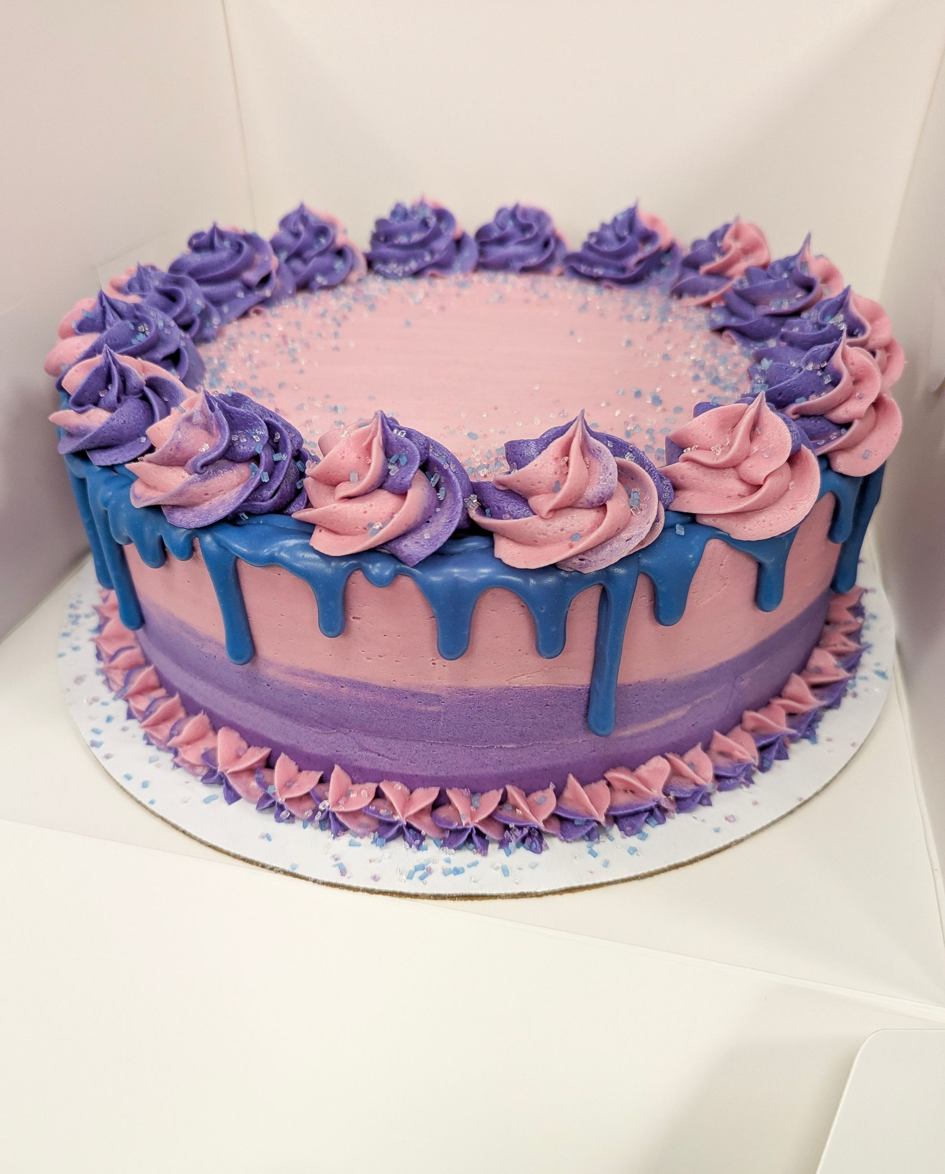 Lavender cake with purple and pink frosting roses and blue drip glaze on a white stand