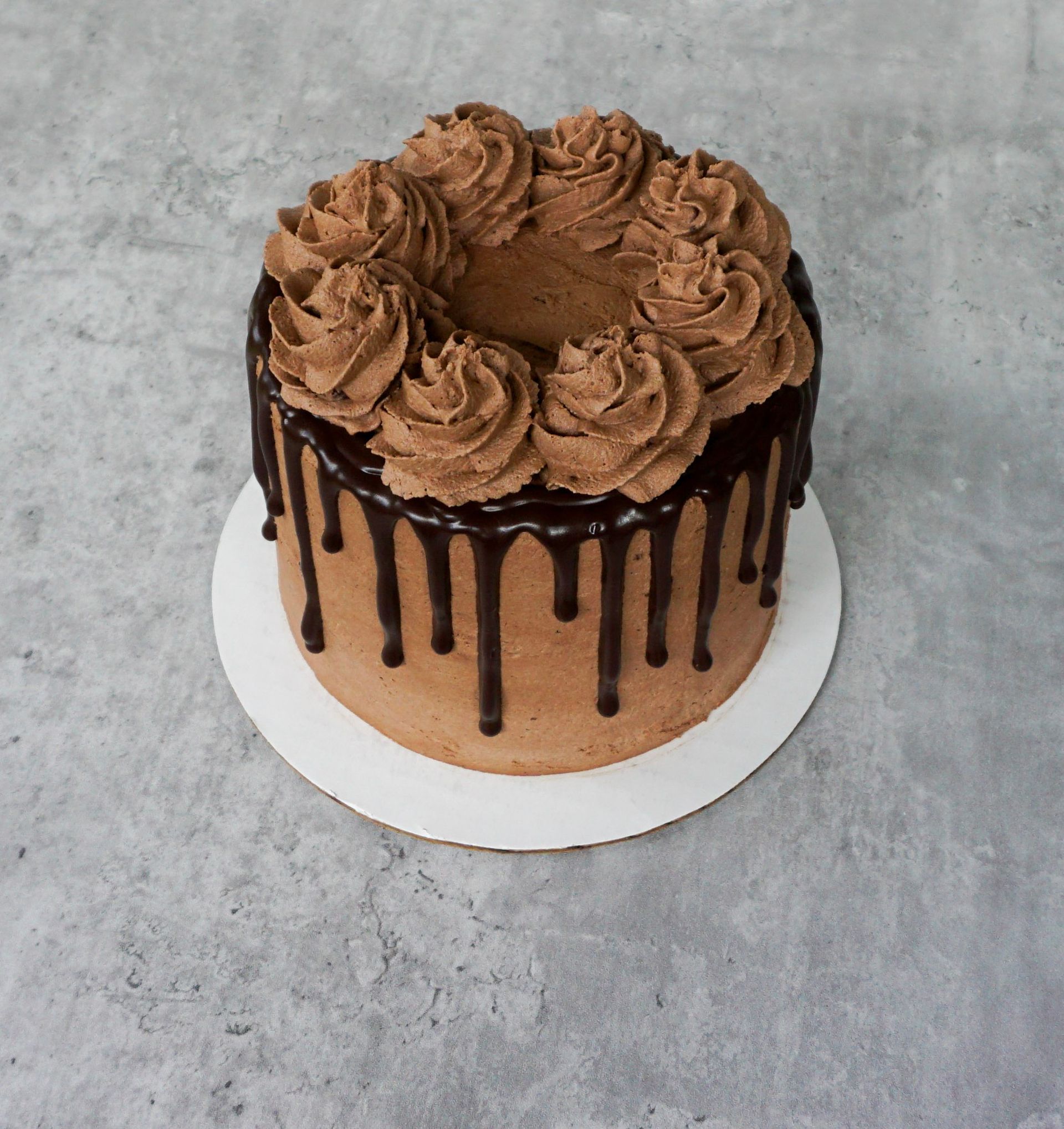 Chocolate drip cake topped with chocolate frosting rosettes on a white cake board