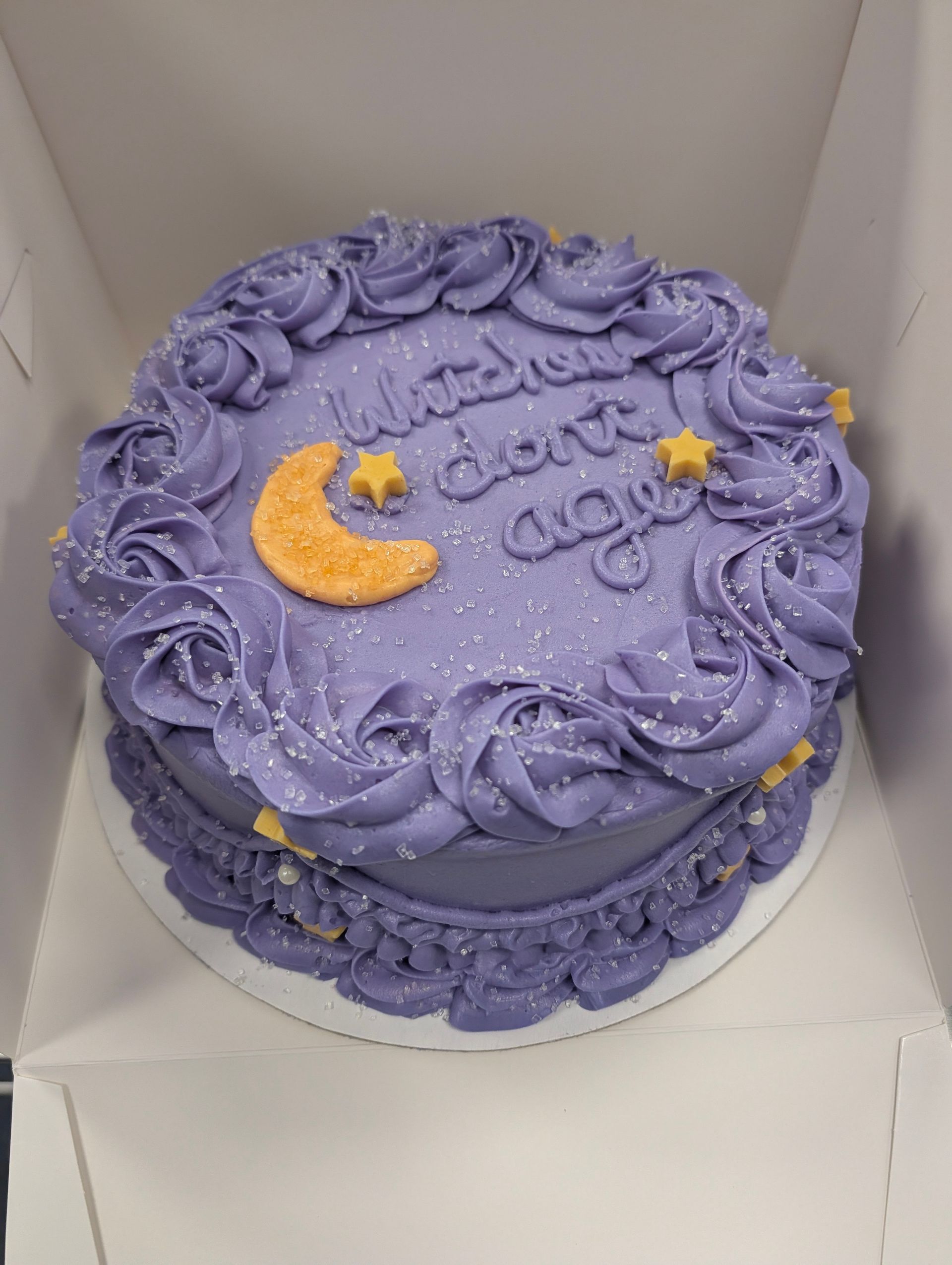 Lavender frosted cake with purple rosettes, a gold crescent moon, and stars in a white box