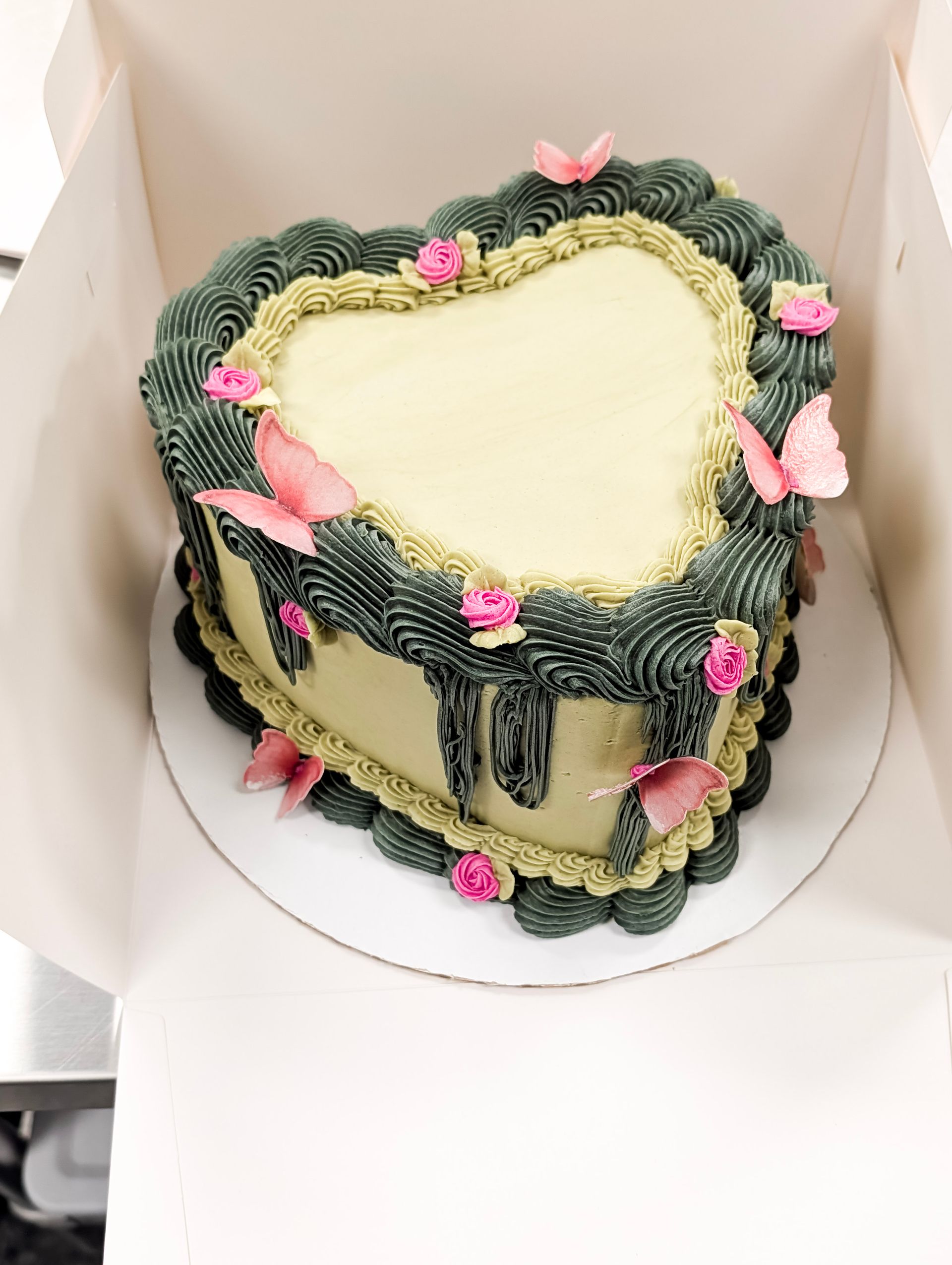 Heart-shaped frosted cake with green piped border and pink flowers in a white box